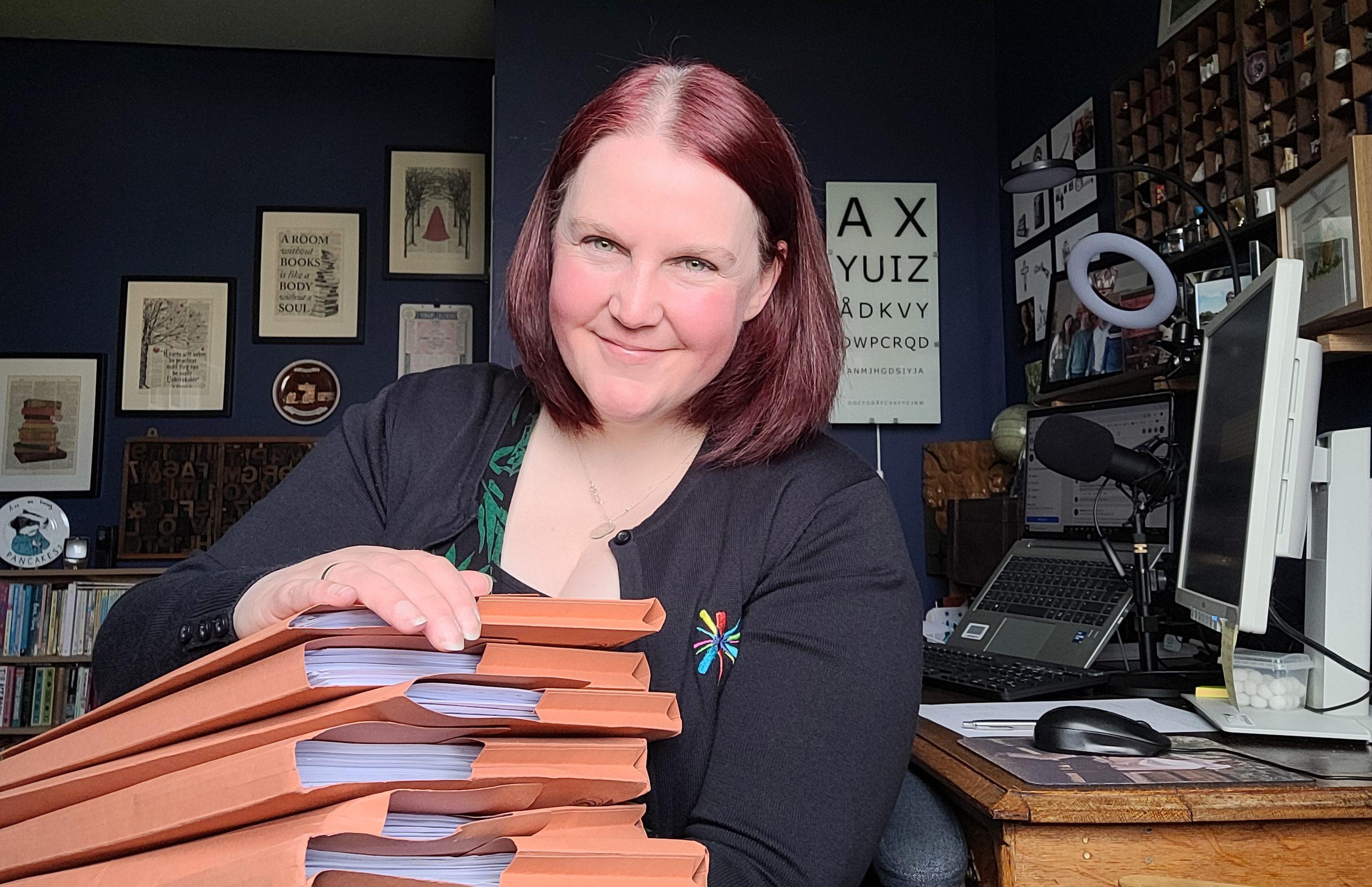 A woman in a black cardigan sitting and holding a pile of orange folders filled with papers.