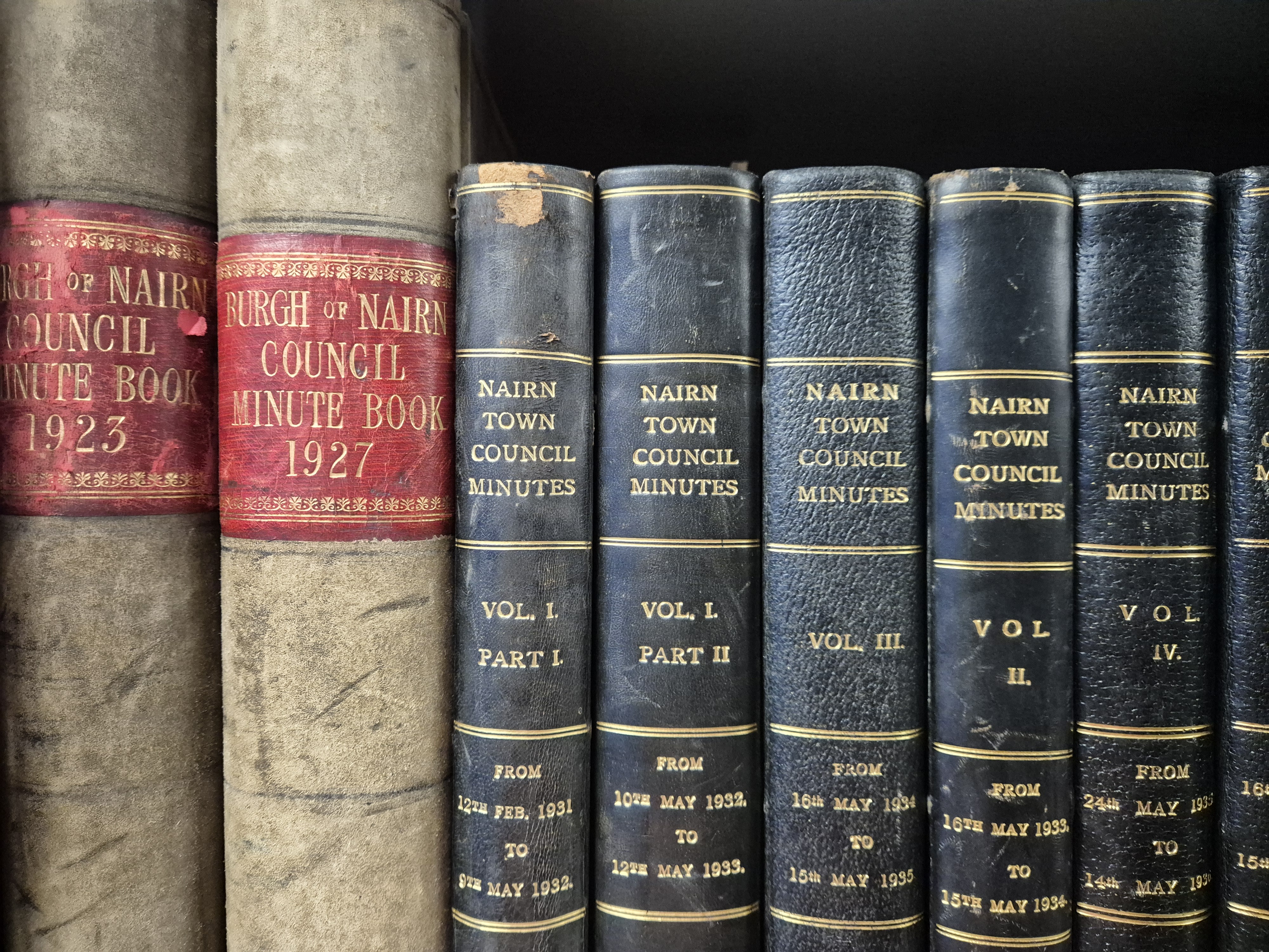 A shelf showing 7 bound volumes (two black and two cream).  Their spines note that they are the minutes of Nairn Town Council