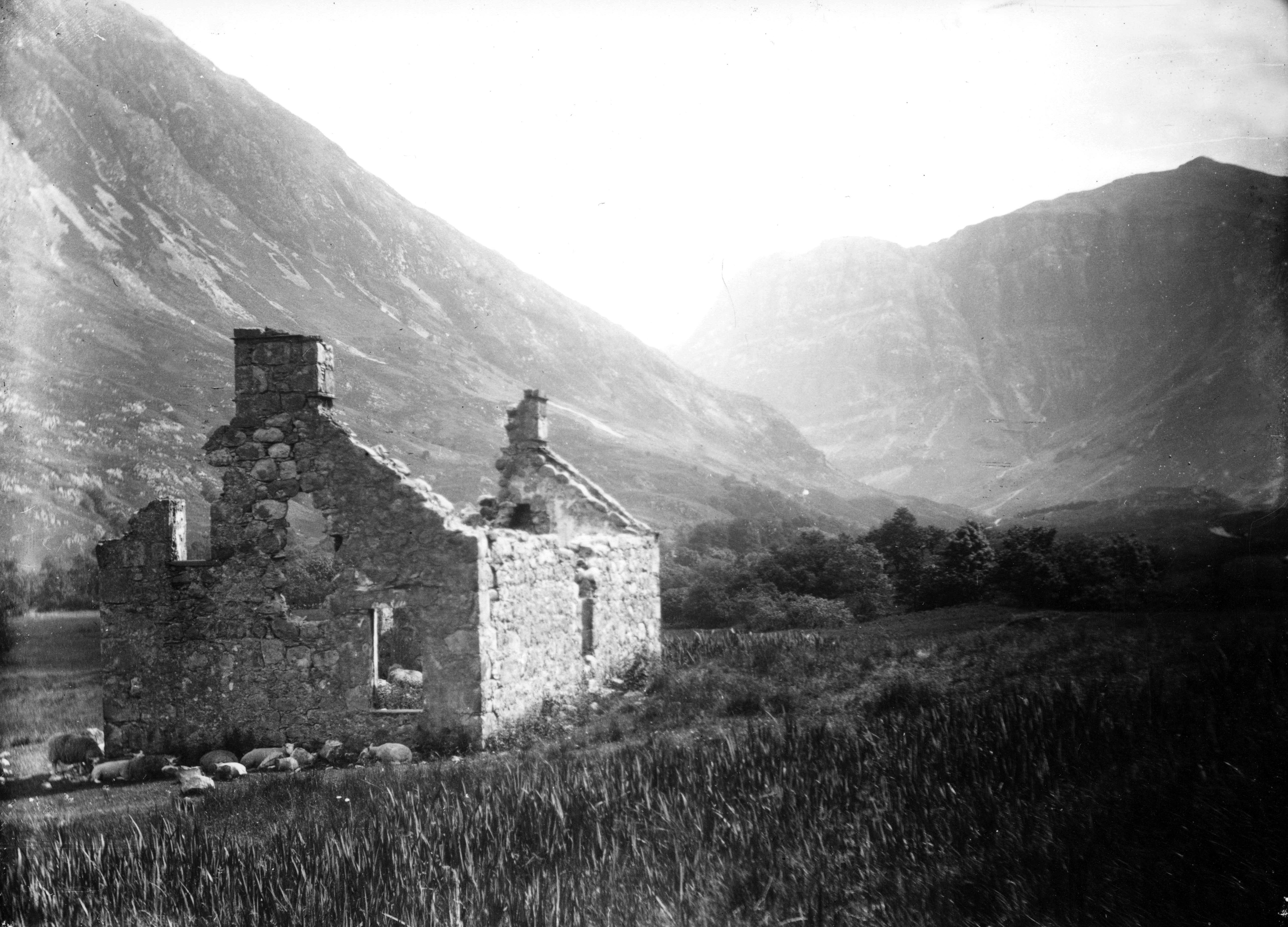 An atmospheric black and white photograph of a ruined stone house against a background of high mountains