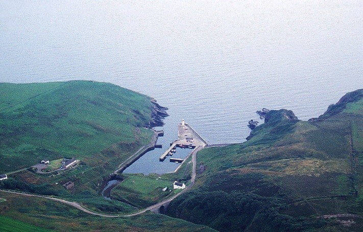 A colour photograph taken from the top of a hill looks down on an empty harbour and quayside