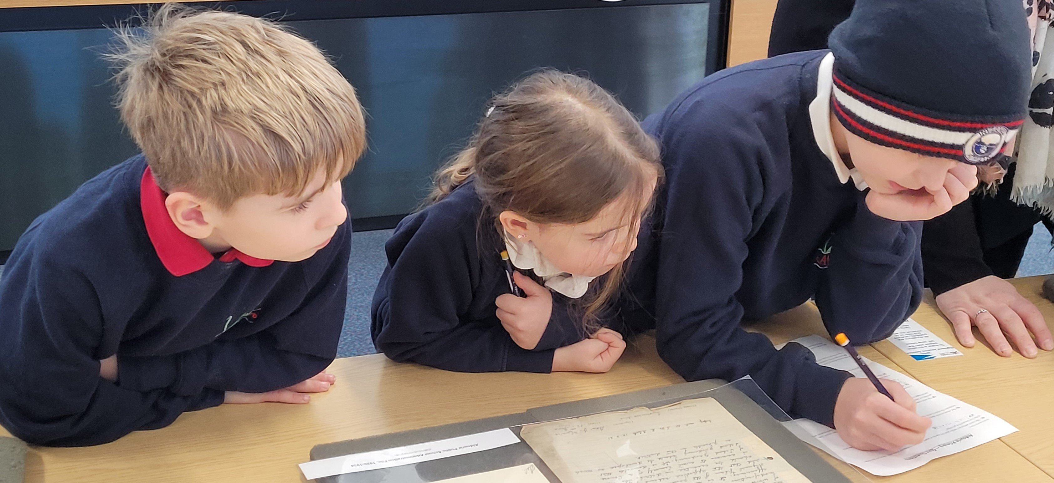 Three children in dark blue school jumpers leaning over a table.  They are looking at a document on a foam rest and making notes on a piece of paper.