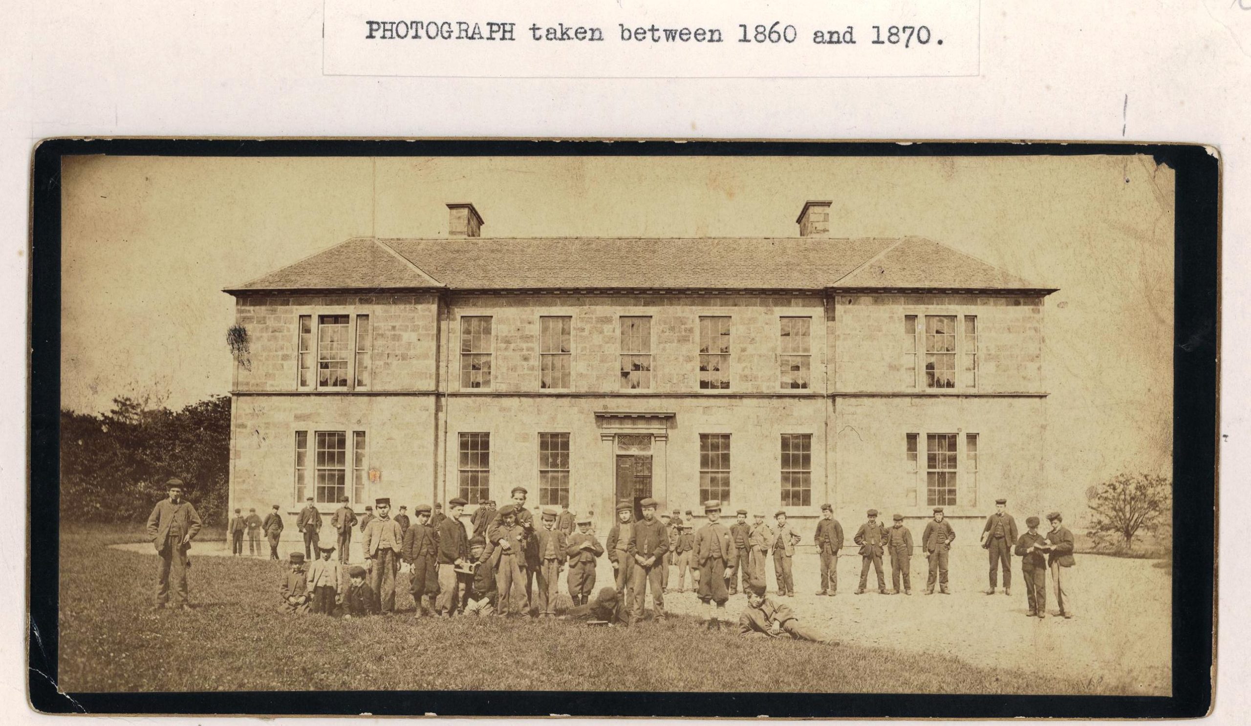 A sepia-coloured photograph of a two-storey school building with children standing and lying in front of it. A typed caption reads 'Photograph taken between 1860 and 1870'