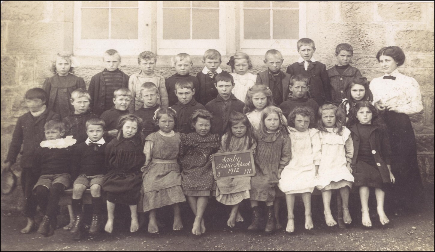 A sepia toned photograph of children sitting and standing in tiers with a teacher to one side.  Many of the pupils have bare feet and one child in the front row is holding a sign which reads 'Embo Public School 1912"