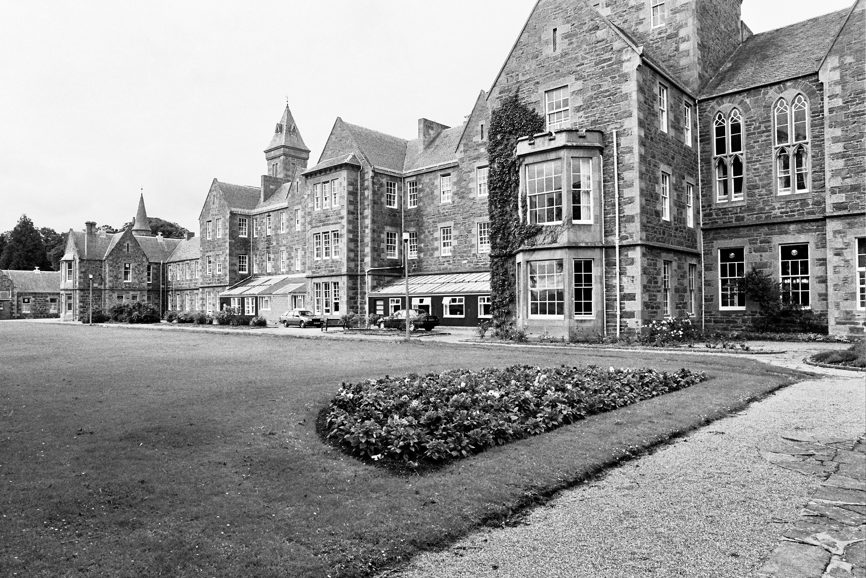 A black and white photograph showing a large and imposing Victorian hospital building with a large lawn and several cars in front