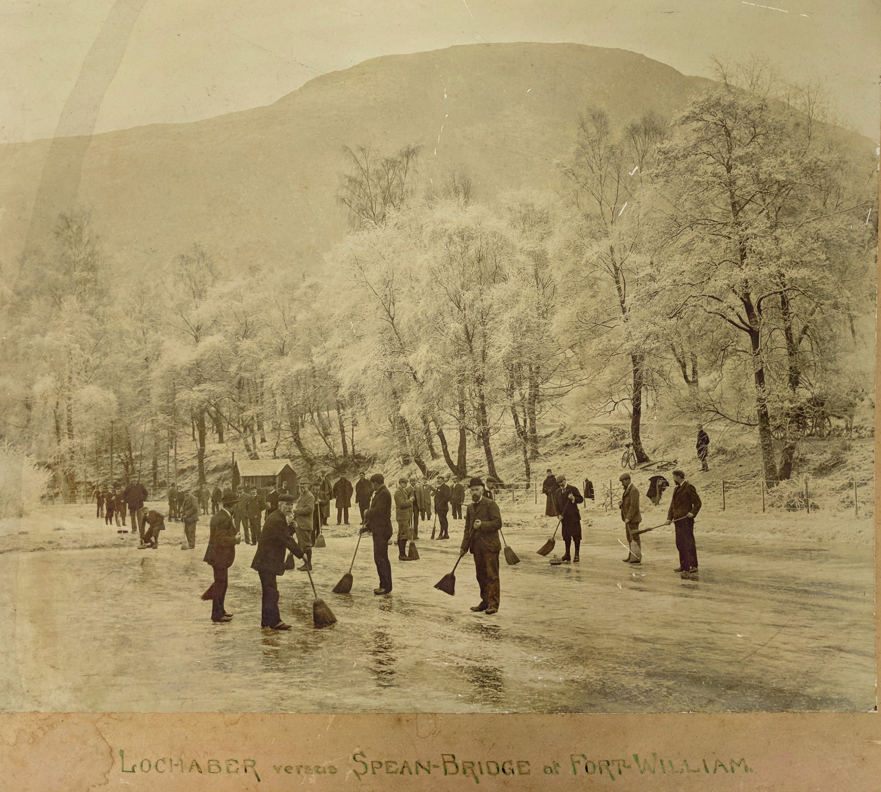A sepia photograph shows men in Victorian attire holding a curling match on a frozen loch.  There is a tree-lined hillside in the background and a caption indicating that the image depicts Lochaber versus Spean Bridge at Fort William