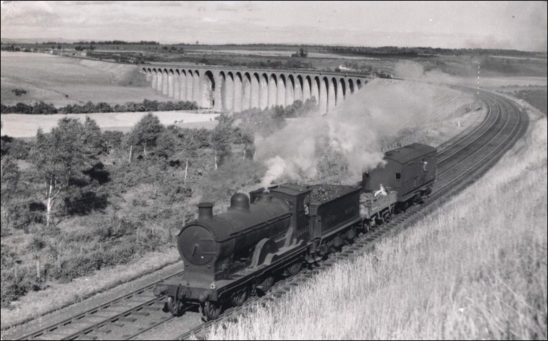 A black and white photograph shows a train engine in the foreground which has just passed over a long sweeping curved viaduct in the background.  There is a rural landscape on either side