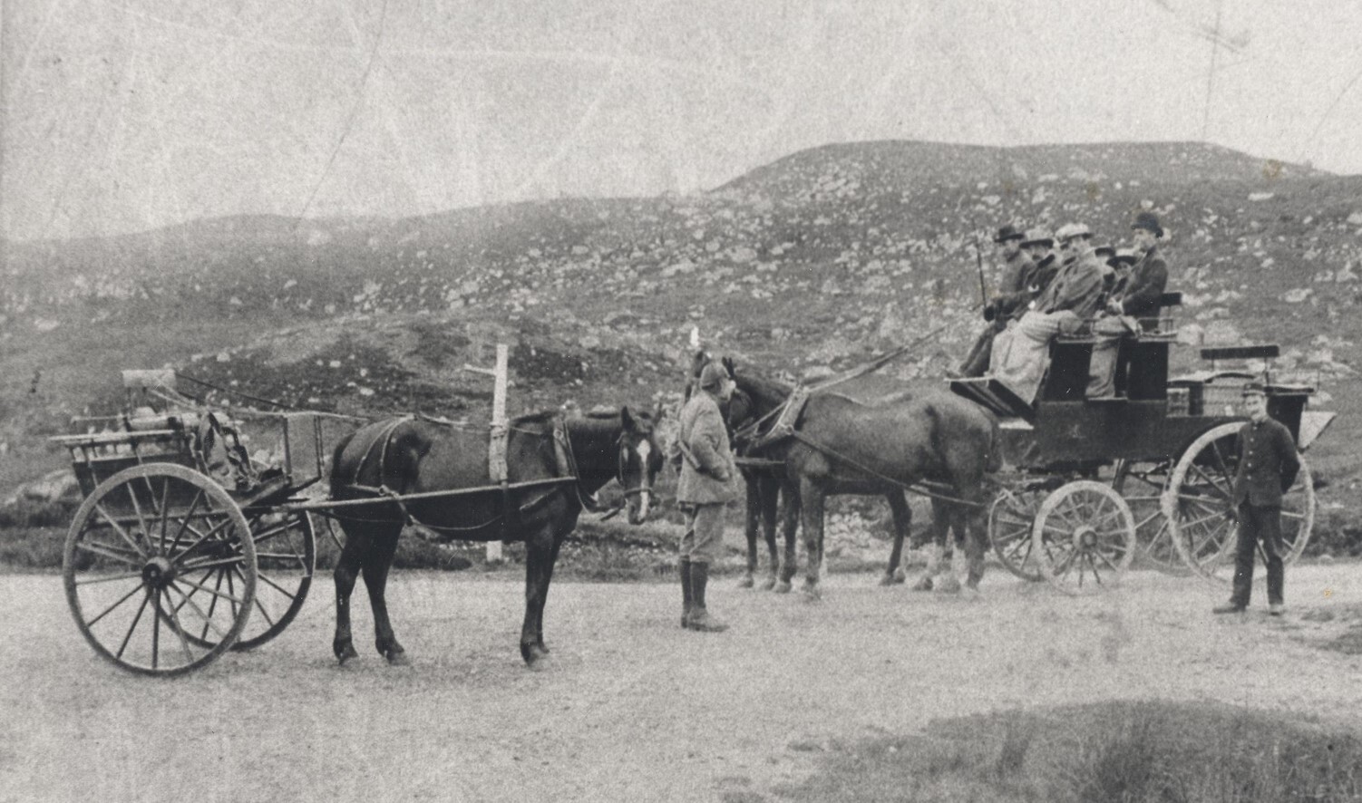 A black and white photograph shows rural landscape in the background with two horse-drawn vehicles in the foreground.  The lefthand one is a small cart with a man standing next to it. The righthand one is a larger vehicle with passengers sitting on top