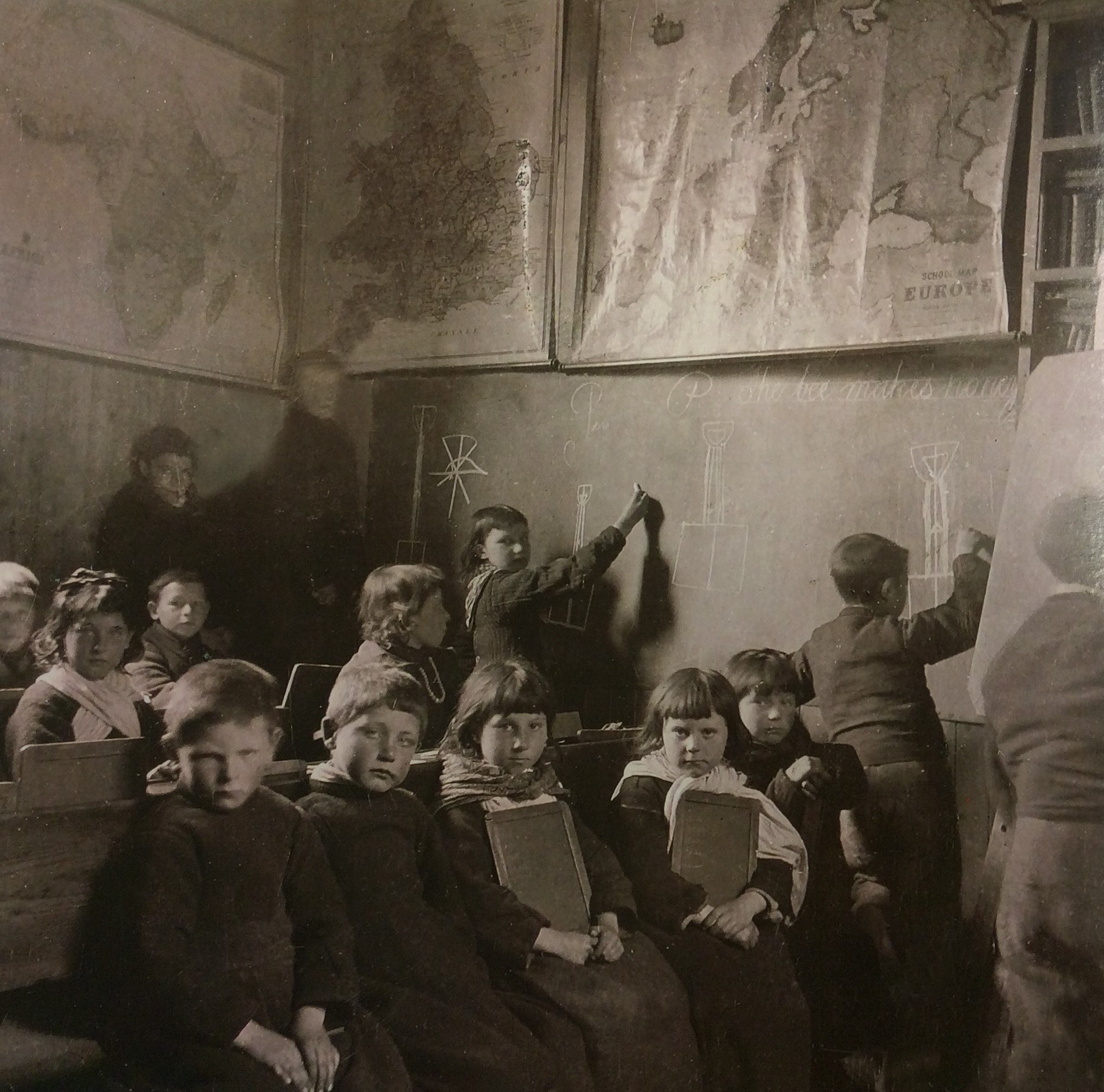 Black and white photograph showing children sitting in lines in a small room.  The wall to the right hand side is being used as a blackboard.  Maps are hanging on the walls.