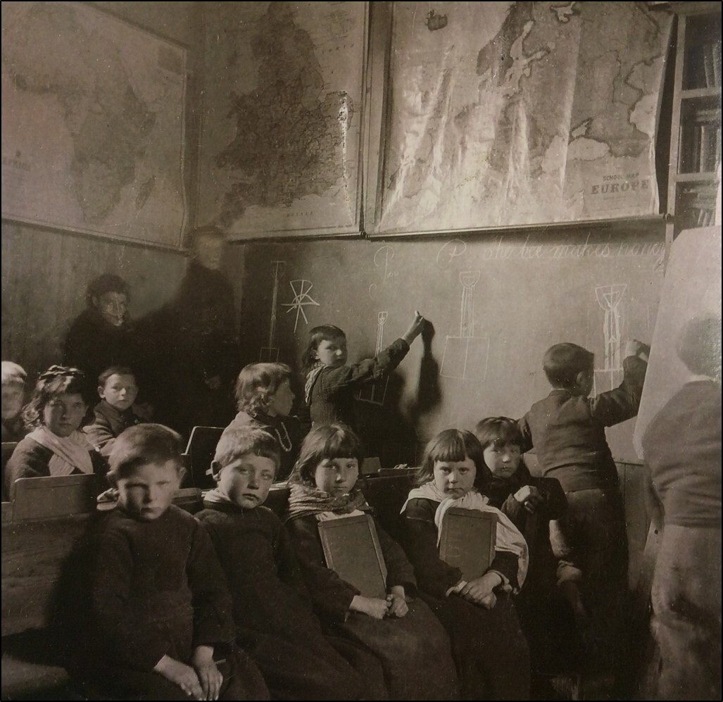 A black and white photograph showing children sitting in rows on benches.  They are holding slates for writing on and some are standing to the right of the photograph writing on a large blackboard.  There are maps hung on the walls