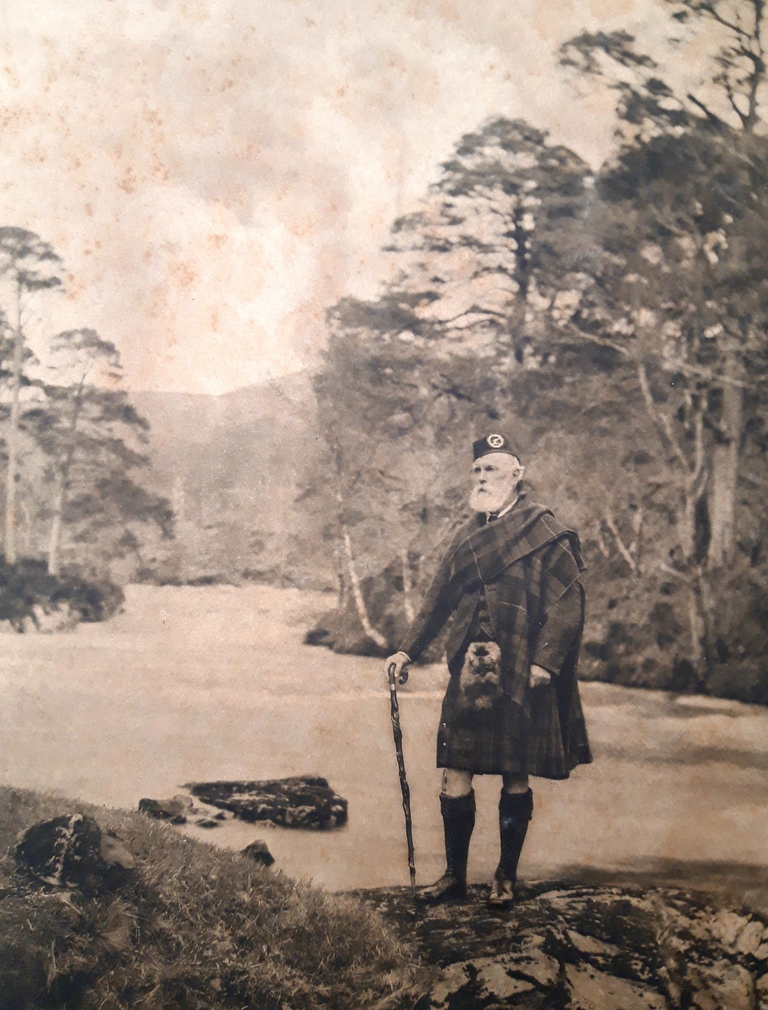 A sepia picture of a man in Highland dress holding a walking stick and facing to his right.  The backdrop shows a river with trees on either side