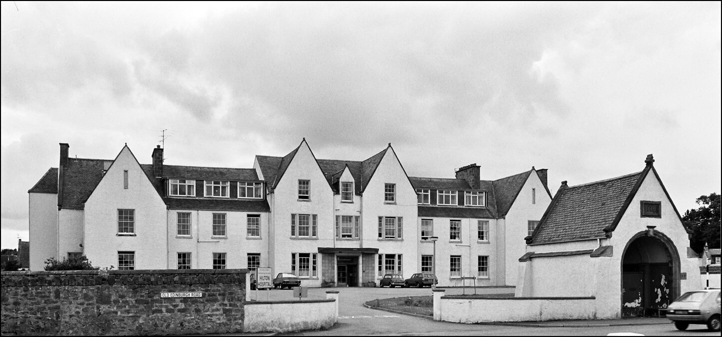 A black and white photograph pf a white, winged, two-storey building with a covered entranceway