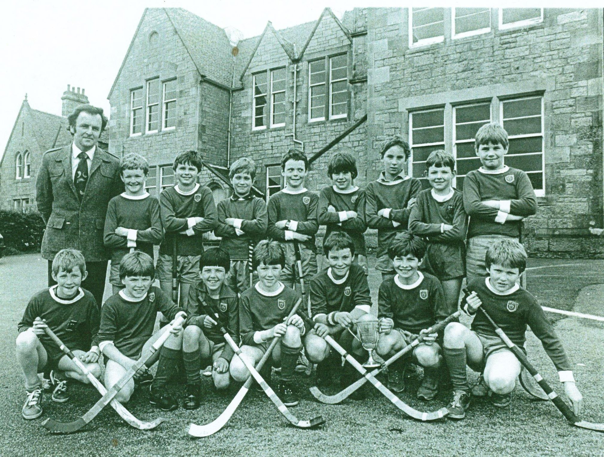 A black and white photograph showing a group of boys with crossed camans (shinty sticks) and a man in front of a large school building