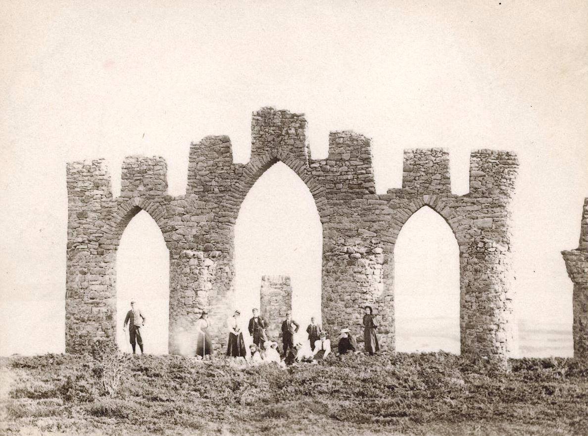 A sepia photograph of a tall three-arched stone monument on a hillside.  A group of people in Victorian dress are gathered in front of it