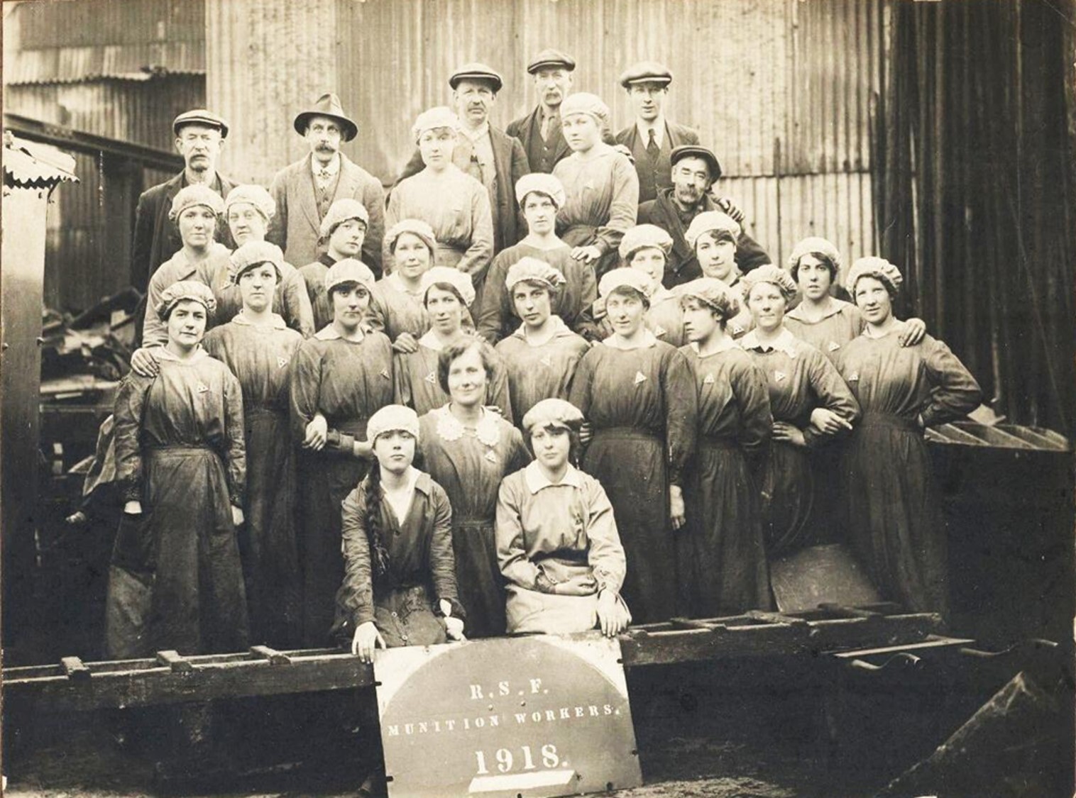 A sepia coloured photograph of a group of women, and some men, in factory clothing and hats, standing in front of a corrugated building.  They each have a small triangular badge on and the two women at the front hold a sign that says 'R.S.F. 1918'