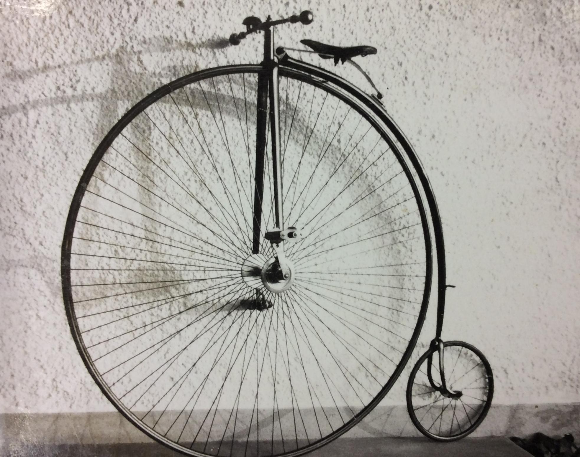 A black and white photograph of a bicycle with one large front wheel and a much small rear wheel