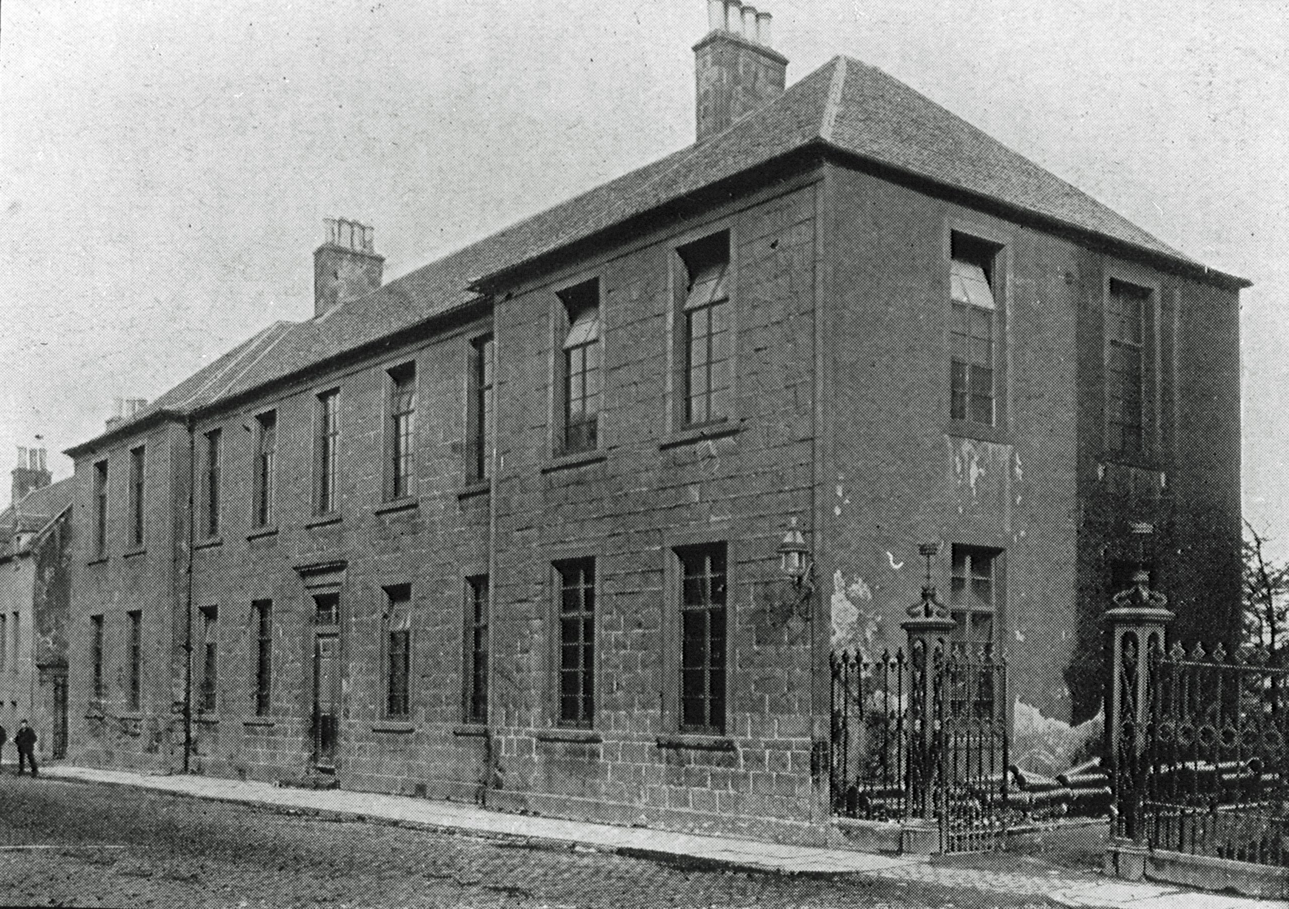 A black and white photograph of a two-storey school building dating from the 1700s. The photograph is taken from an angle which shows the the front and one end of the building.