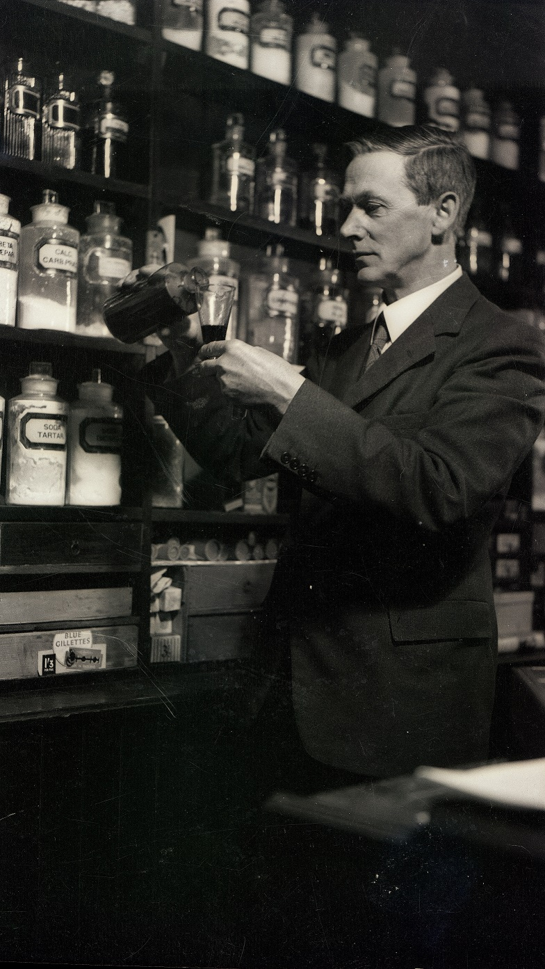 A black and white photograph of an old-fashioned chemist's shop with numerous glass bottles on shelves.  A man on the right side of the photograph is pouring liquid from one to another.