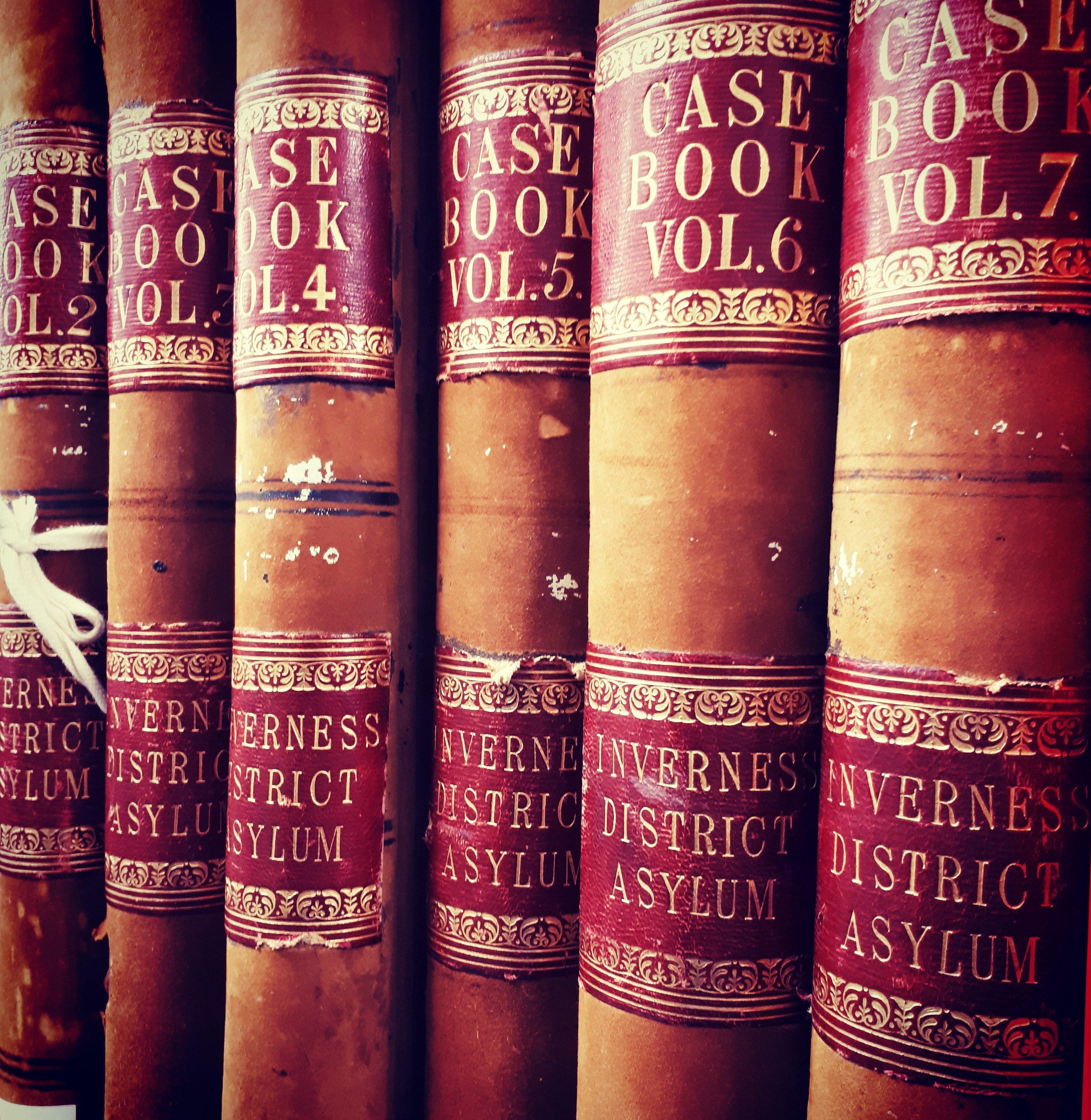A richly coloured image of six large leather-bound volumes with red labels pronouncing them as case books from Inverness District Asylum