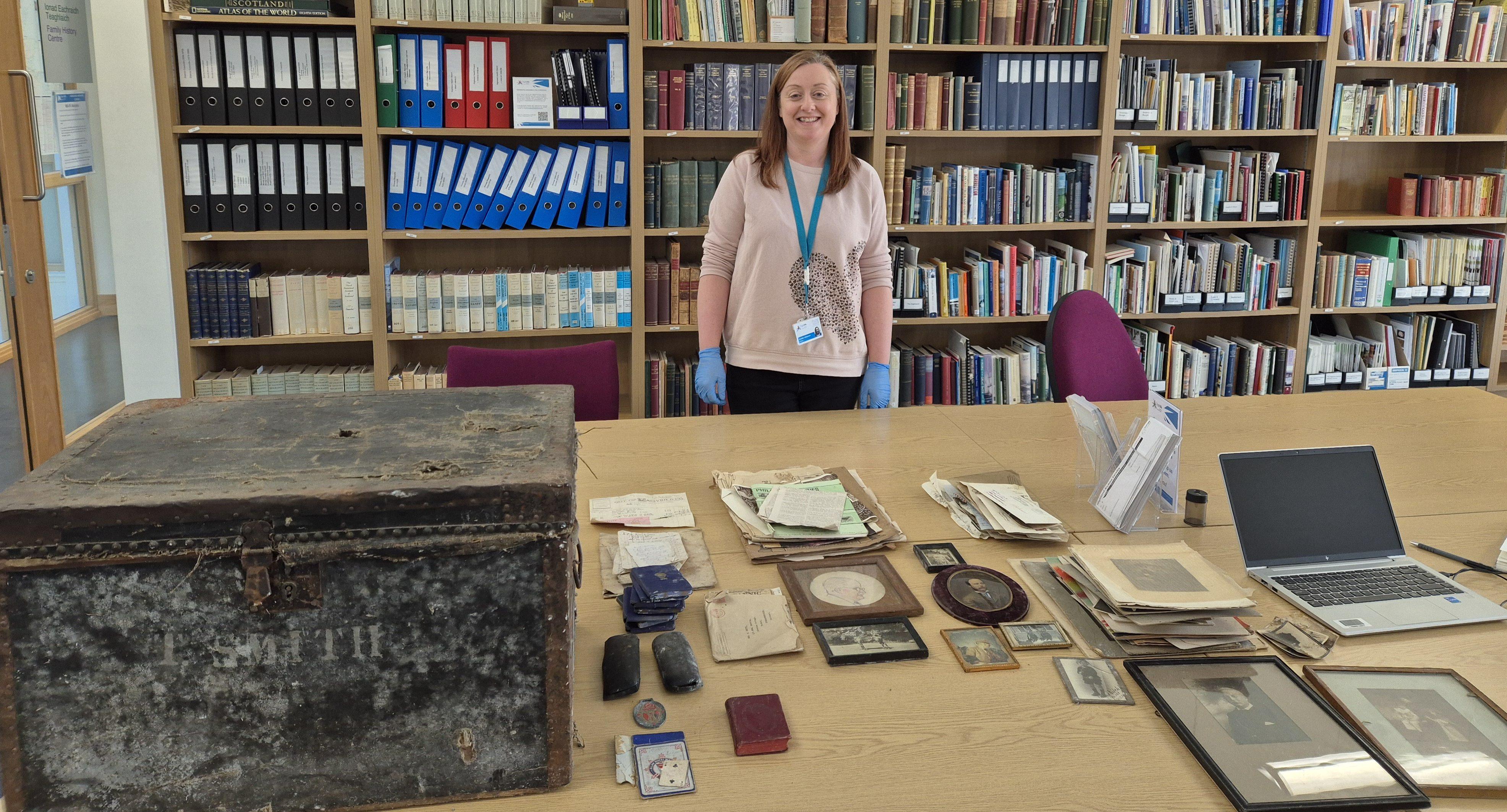 A woman in a pink top behind a table.  The table holds a range of historic documents and photographs, a laptop, and an old wooden chest.