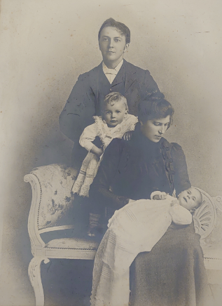 Sepia photograph showing a woman sitting on a chair holding an infant.  A toddler stands behind her on the chair and a man is standing at the back.