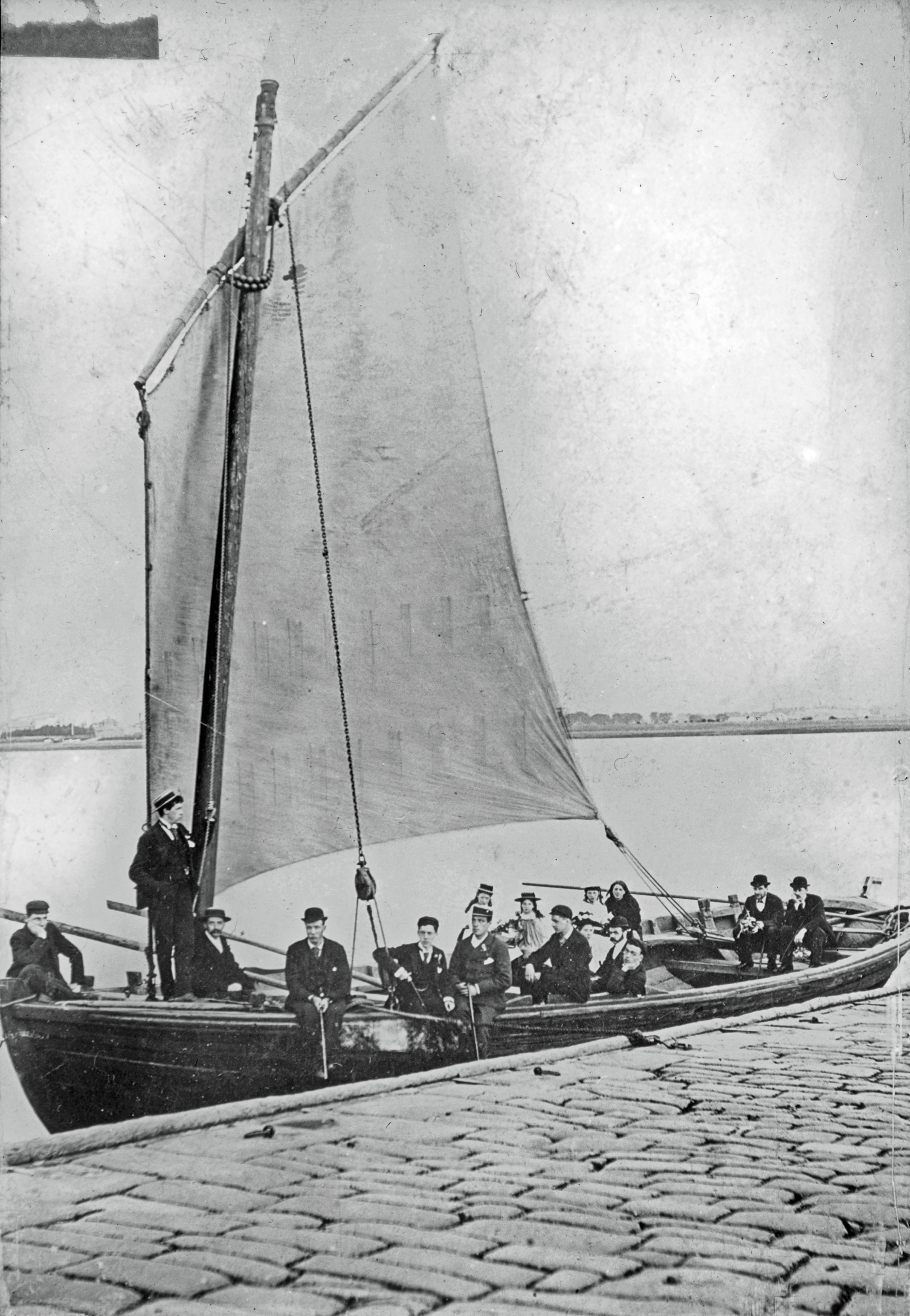 A black and white photograph shows a large sailing boat with its sail unfurled drawn up to a quayside.  There are around 15 people on board in Victorian dress.
