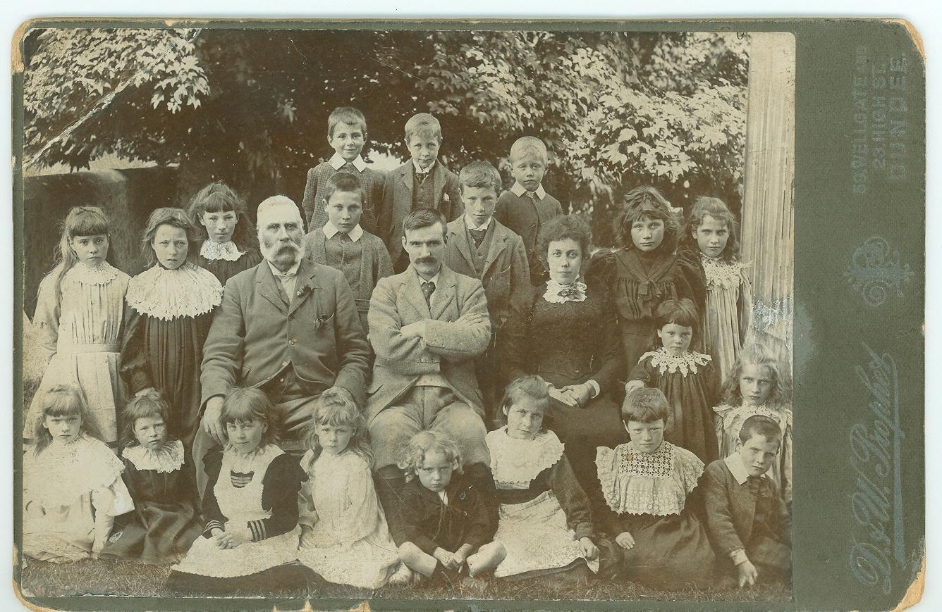 A black and white photograph adhered onto green card produced by D&W Prophet, Dundee.  It shows two men and a woman seated and surrounded by a large group of children.  All are in Victorian dress