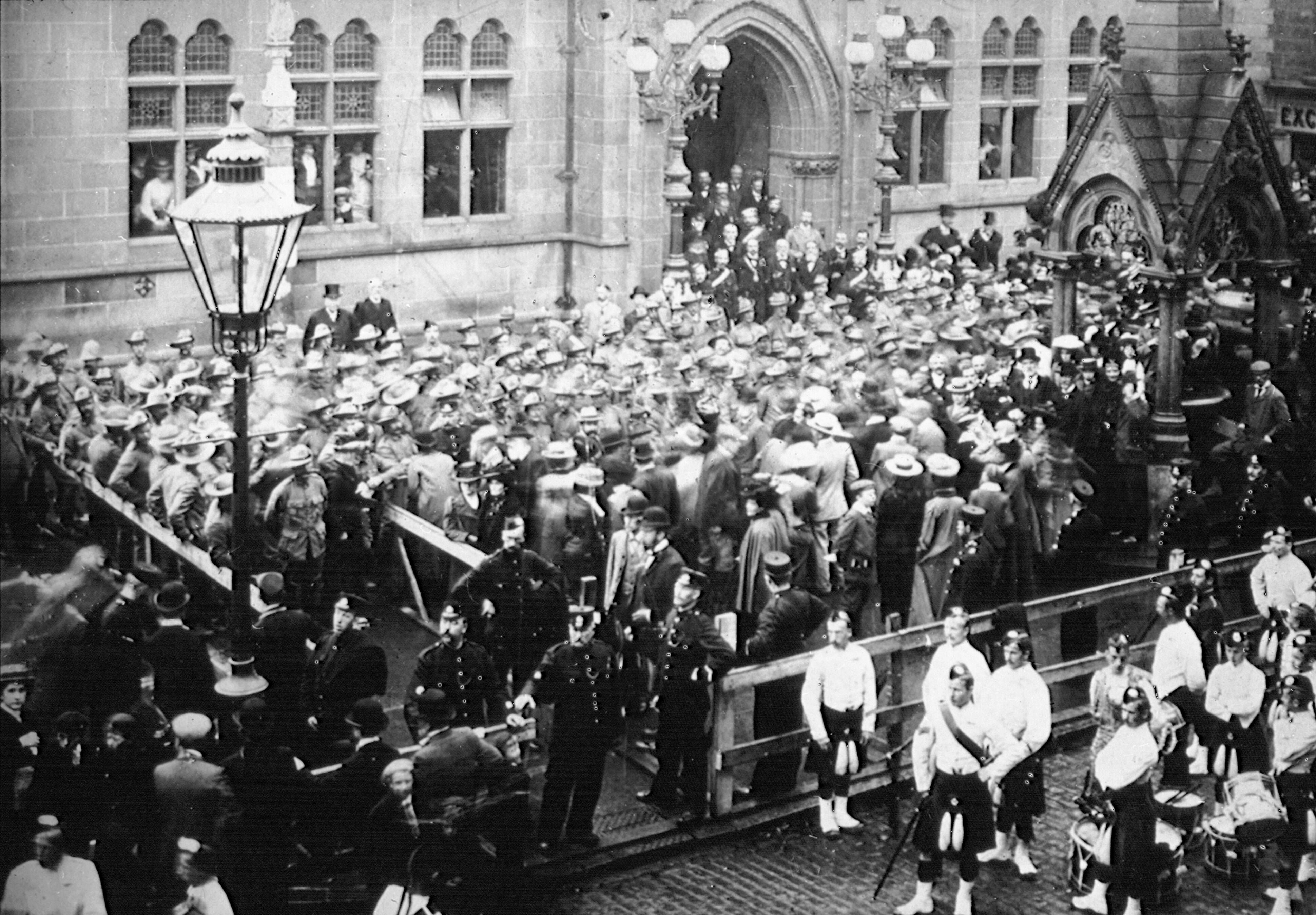 A black and white photograph showing a very crowded scene outside a large building (Inverness Town House).  Men in kilt uniform are visible in the front right of the picture/