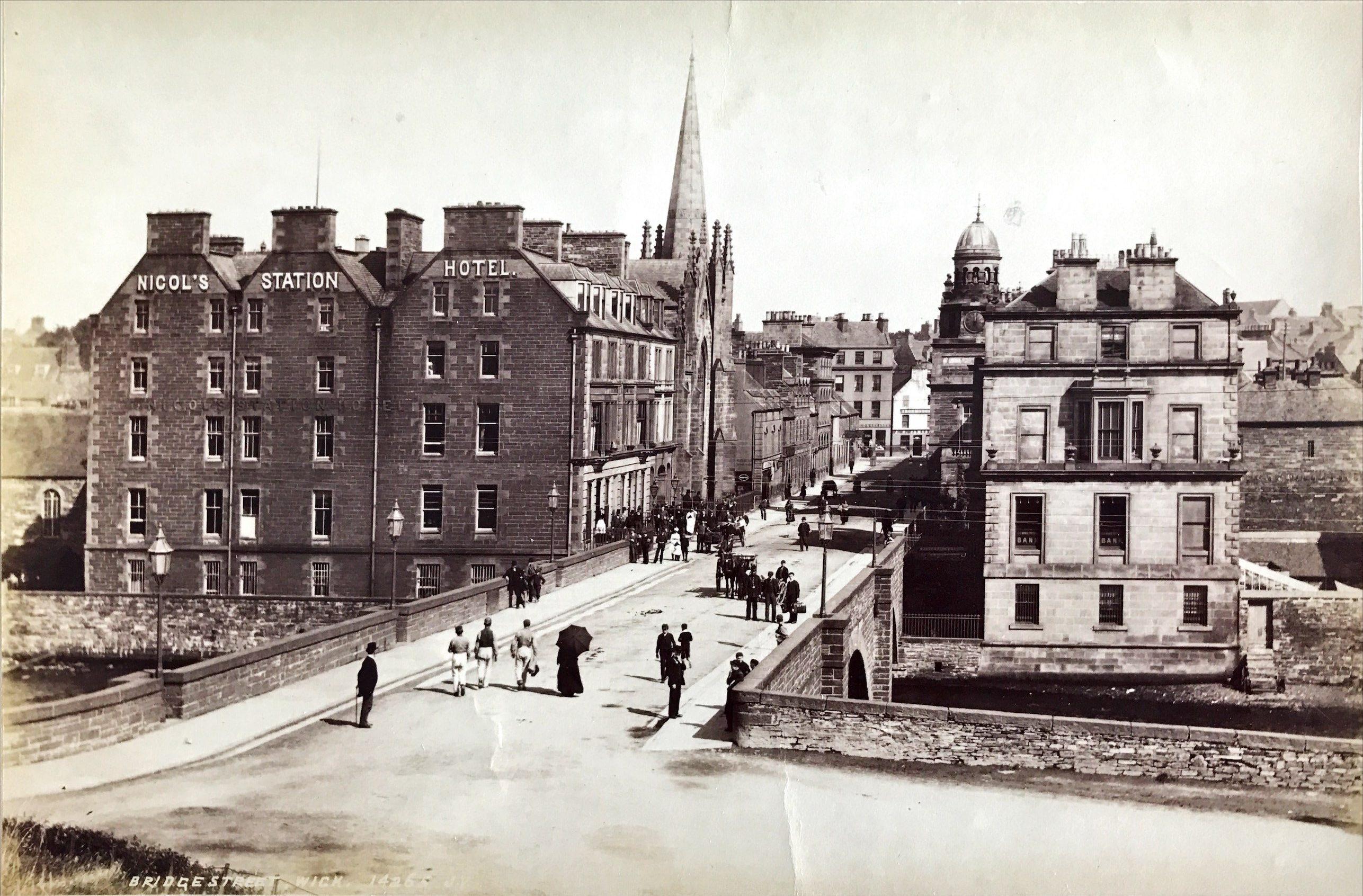 A black and white photograph showing a street scene.  In the centre of the picture people cross a bridge.  The other side of the bridge has buildings on both sides. A building on the left has a sign reading 'Nicol's Station Hotel'