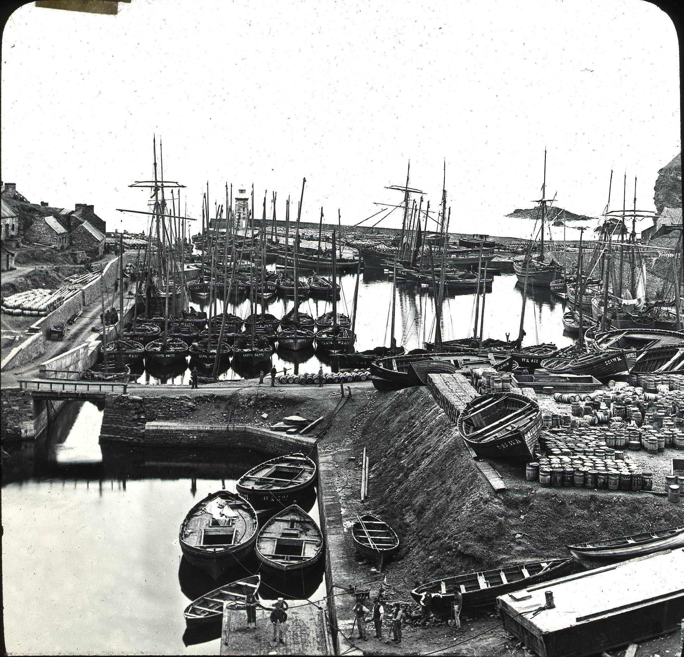 A black and white photograph showing a small harbour full of sailing boats.  To the right hand side a quayside is stacked with barrels