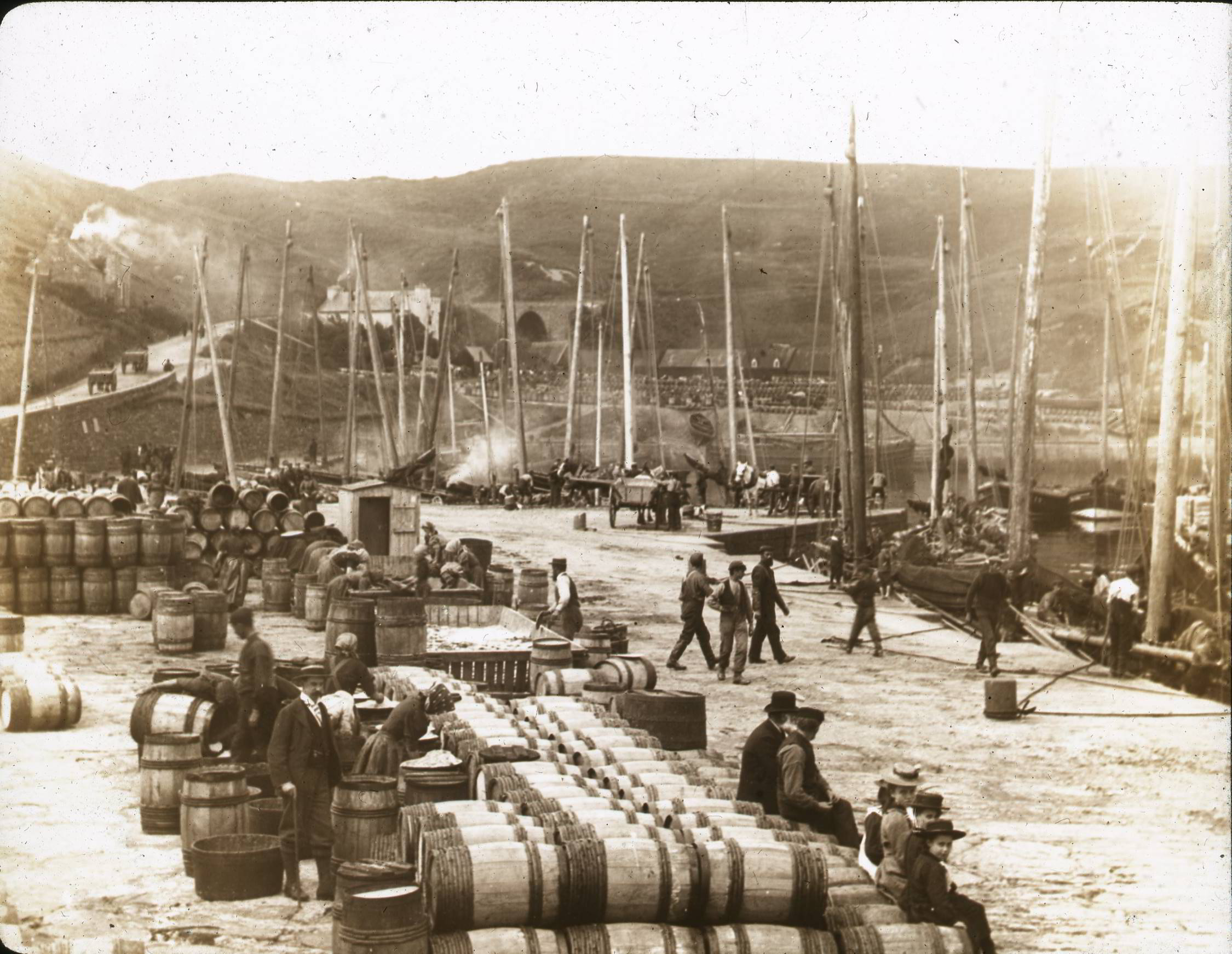 A sepia photo shows a a busy quayside and the hilly landscape beyond.  The quay is stacked with hundreds of barrels and people in Victorian dress can be seen