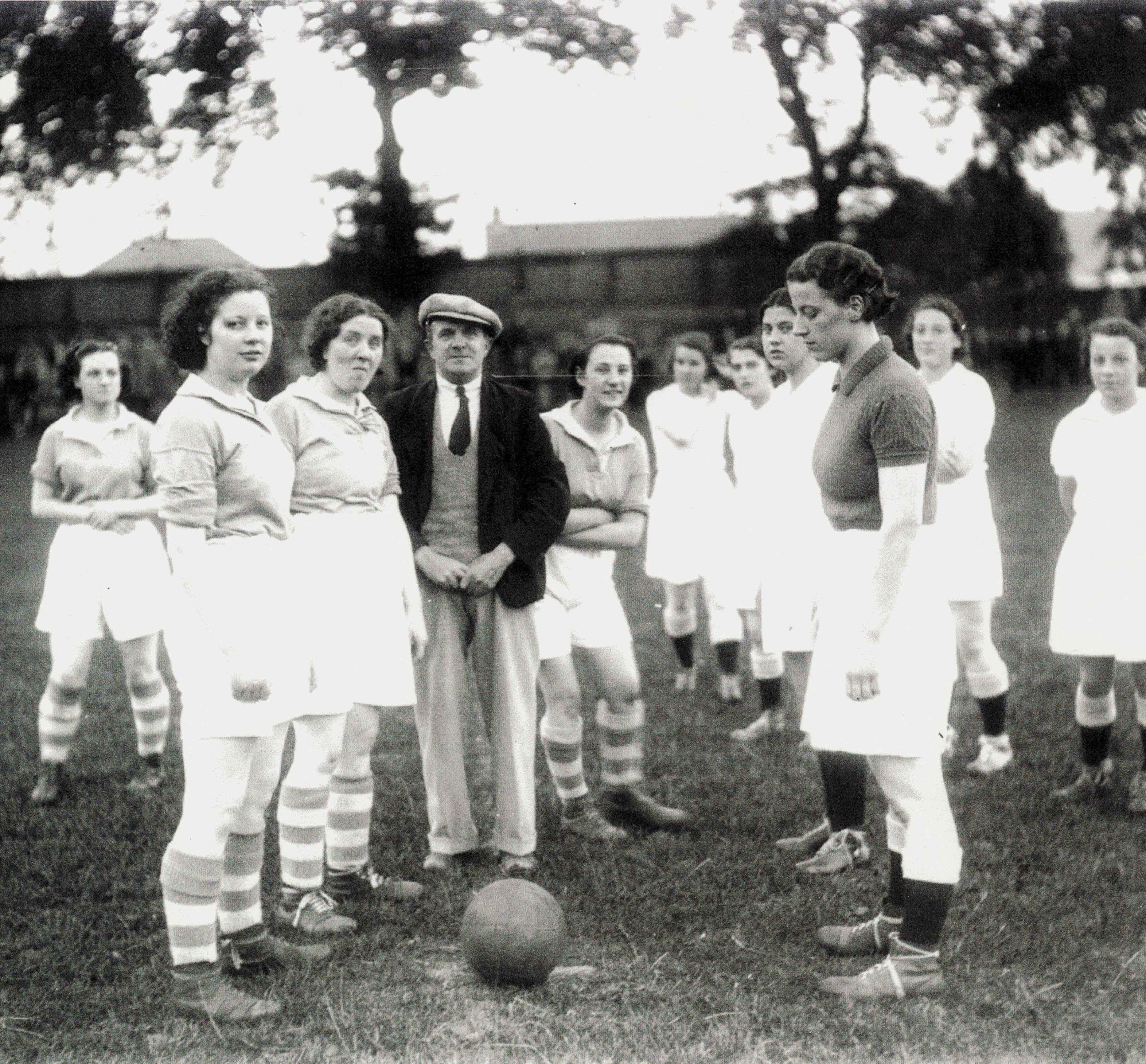 A black and white photograph showing a two teams of women in football kits of the 1930s. A ball lies at their feet and a man in a suit is in the centre of the photograph.