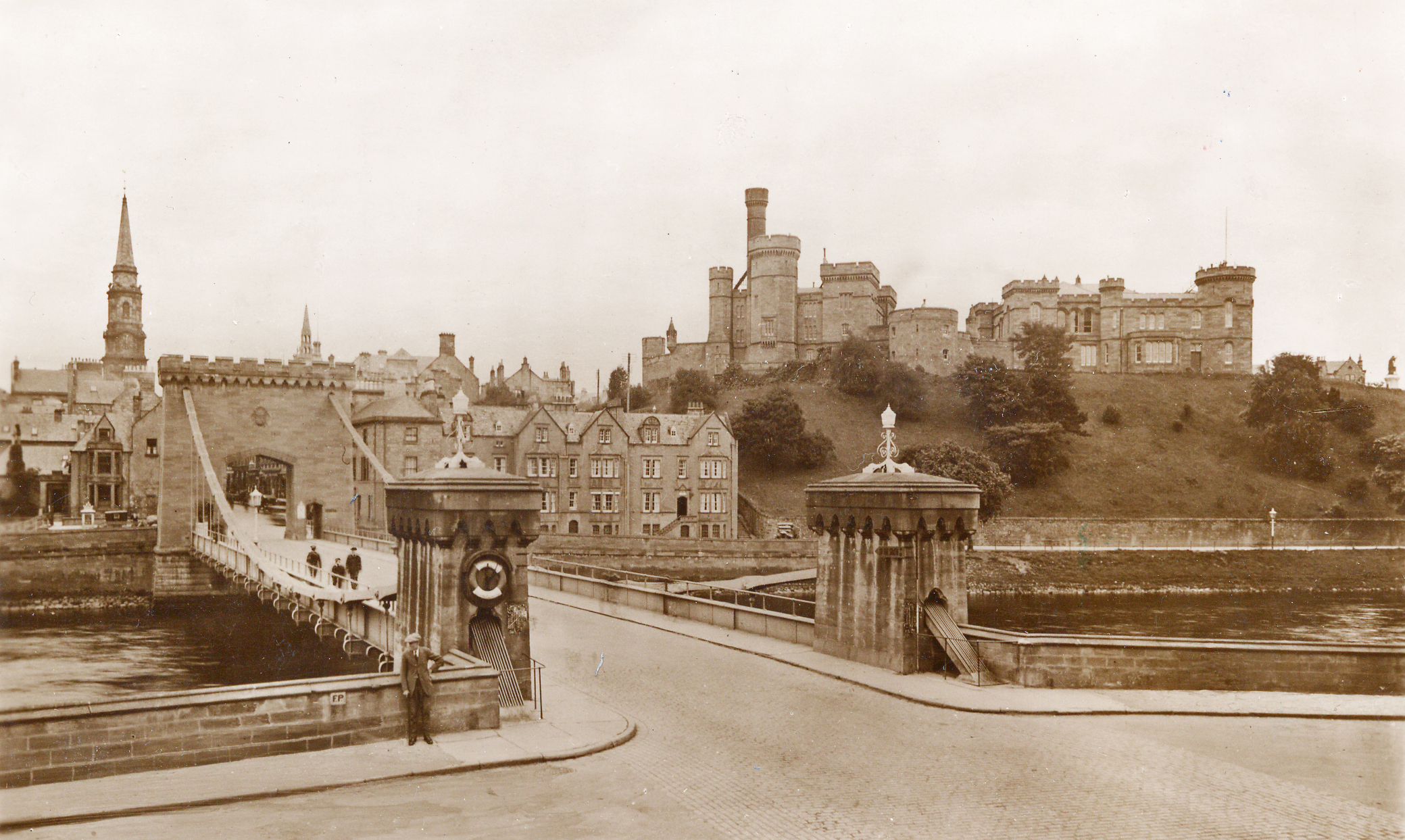 A sepia coloured photograph with a bridge and road in the foreground and a 19th century castle on a hill in the background