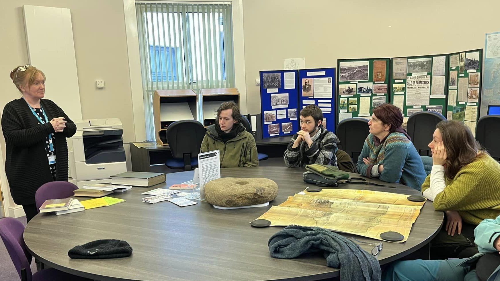 A group of people at a round table in a round room.  One person is standing speaking to the class.