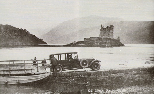 Black and white photograph showing a vintage car in the foreground with the ruined Eilean Donan castle behind
