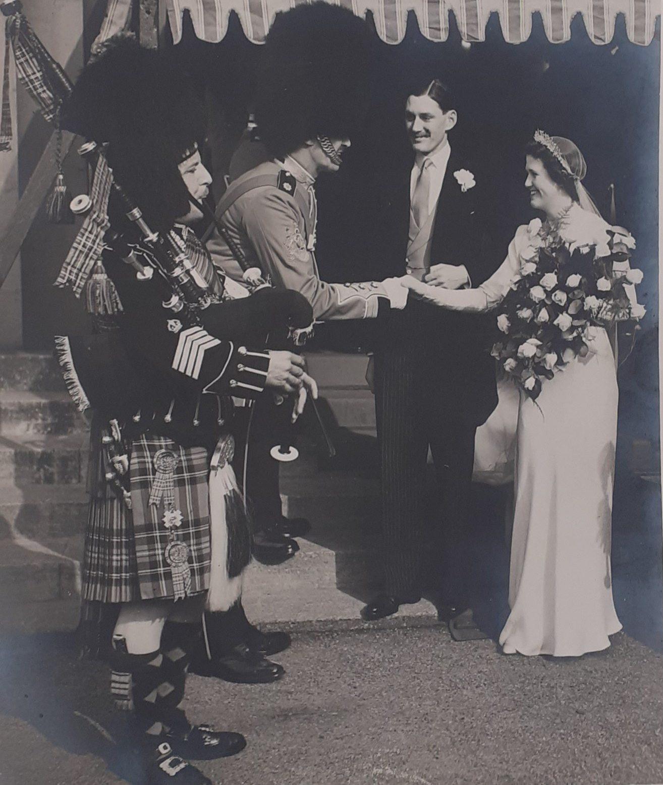 Black and white photograph showing a bride and groom in 1930s fashion.  The bride shakes hands with piper in Highland dress.