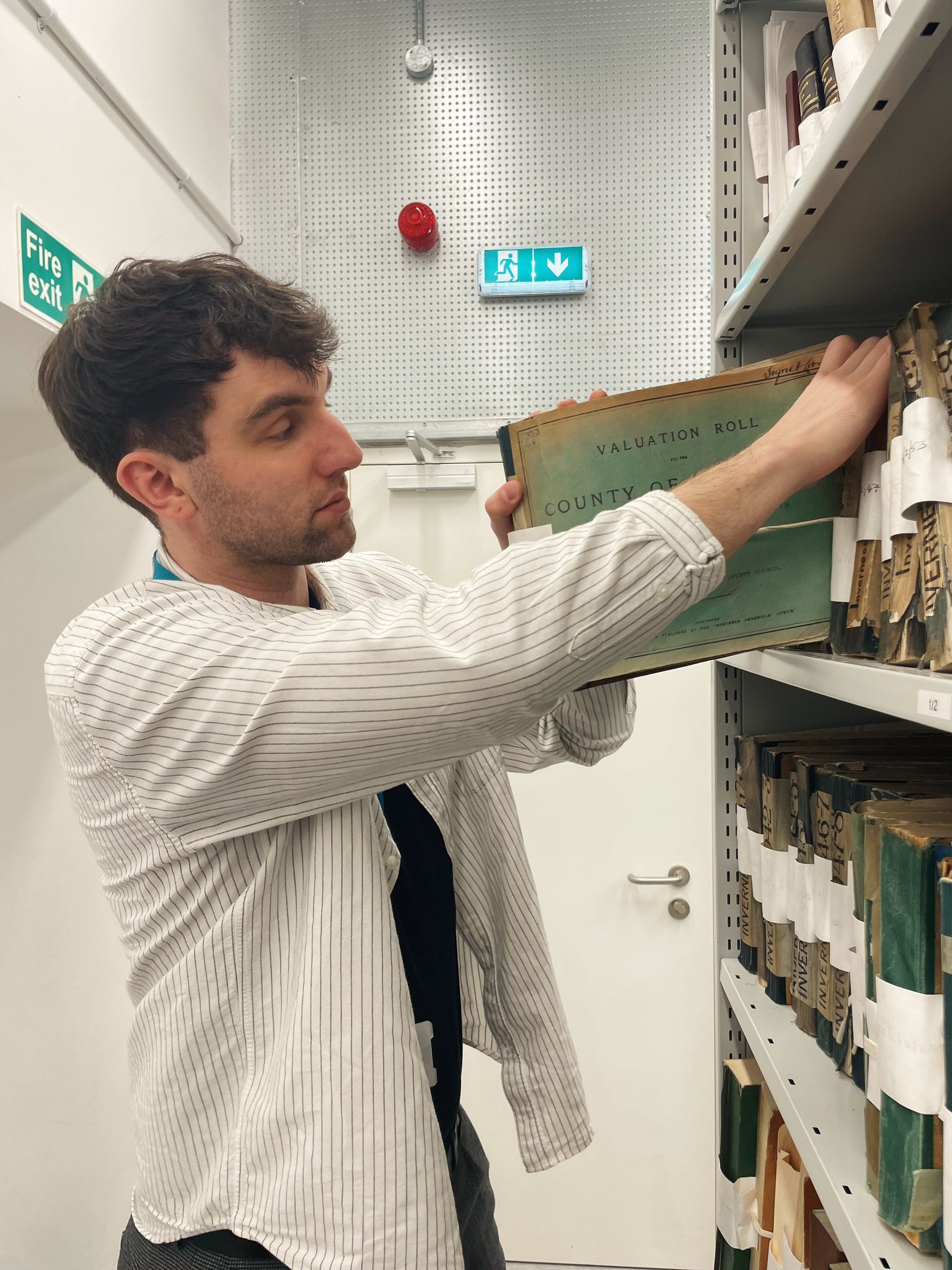 A person wearing a light striped shirt and a blue lanyard is standing beside a shelving unit in an archive storage area, removing a large green-bound volume labeled “Valuation Roll” from the shelf. The shelves are filled with similar large books wrapped in protective covers. A white door and fire exit signs are visible in the background.