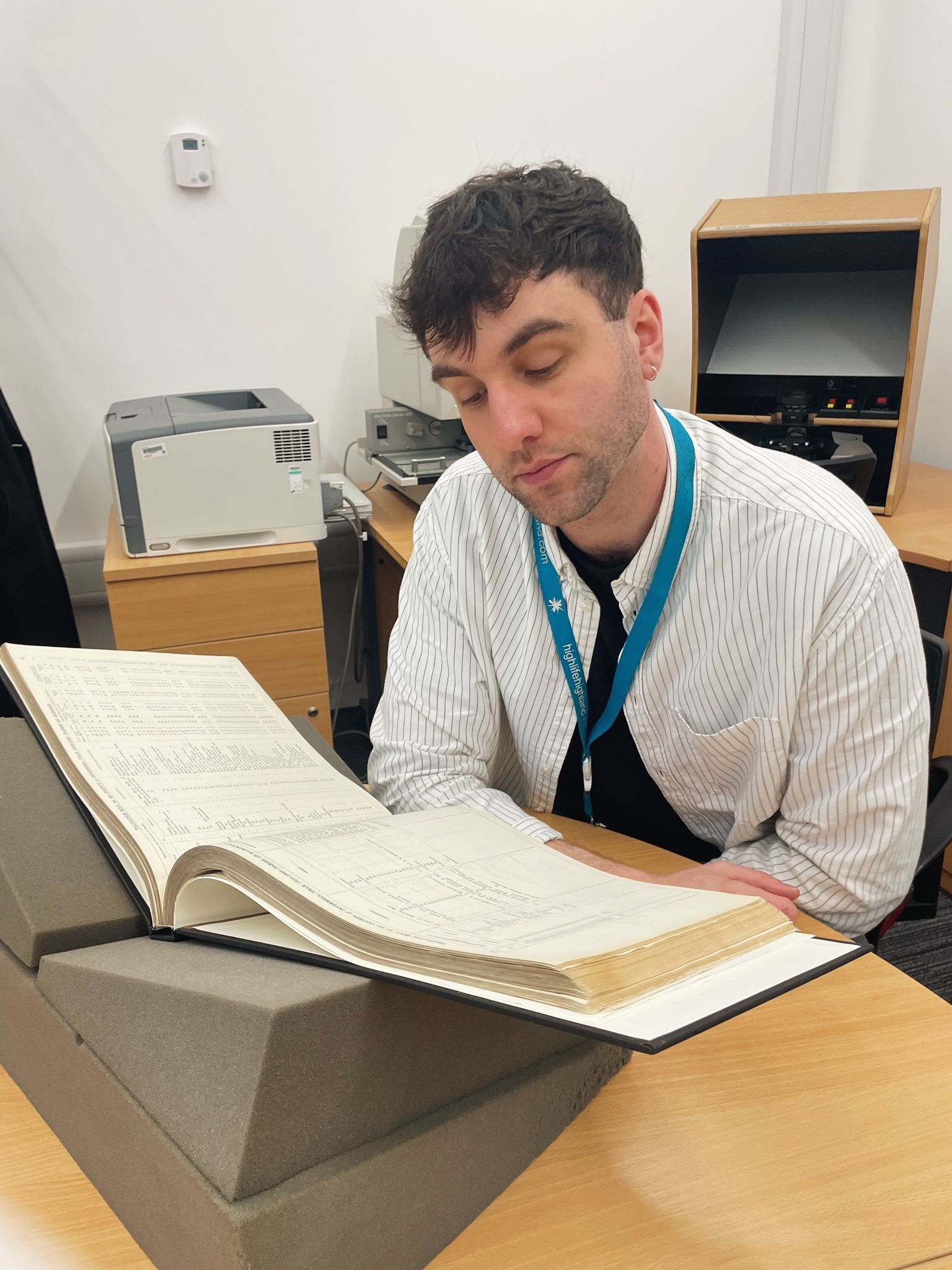 A man wearing a white striped shirt and a blue lanyard is seated at a wooden desk in an archive or research room. In front of them is a large open ledger or archival book resting on a foam book cradle. Behind the desk, there is office equipment including a printer and a microfilm reader.