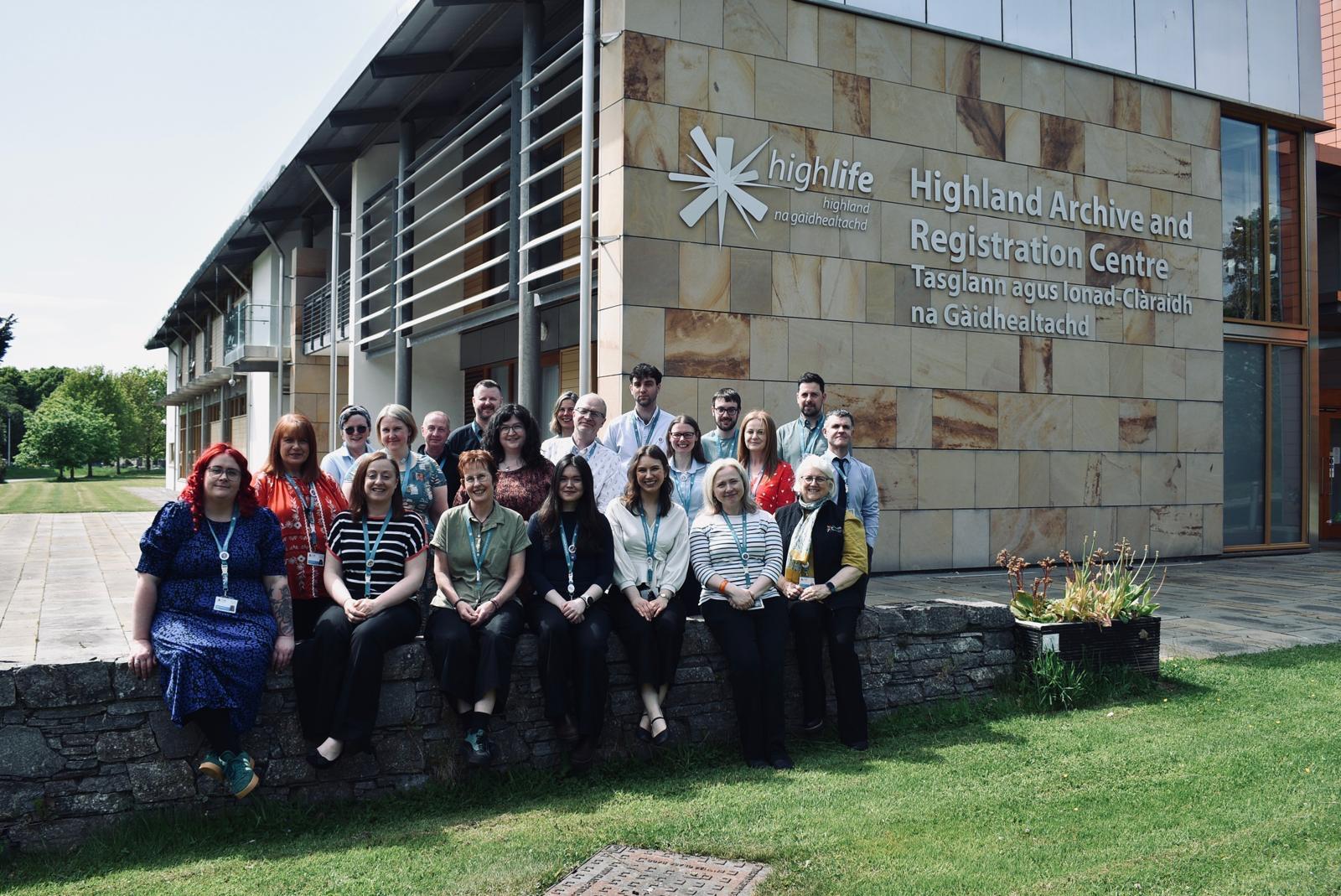 Group photograph of Highland Archive Service staff gathered outside the Highland Archive and Registration Centre in Inverness. Team members are seated and standing in front of the building, with the High Life Highland logo and centre name displayed on the stone-clad exterior