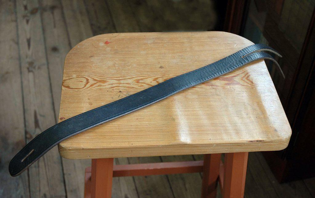A Lochgelly tawse, a thick leather strap with two tails, resting diagonally on a wooden stool with a visible wood grain surface.