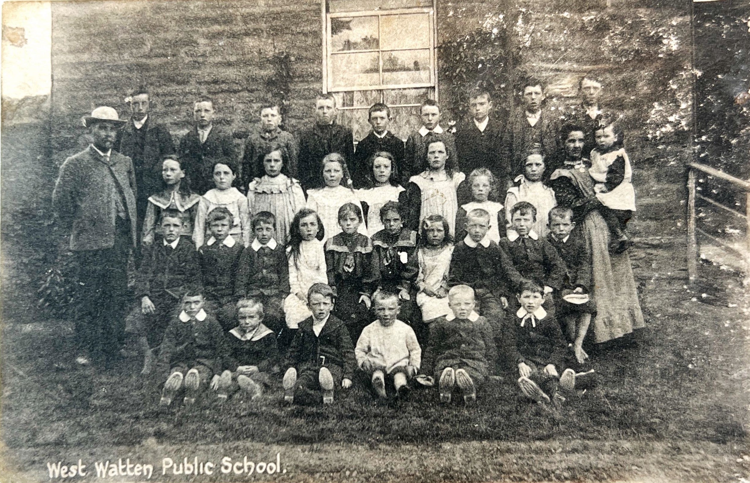 Group photo of children and adults outside West Watten Public School, posed in rows against a stone wall. Date 1910.