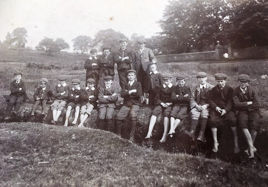 A black-and-white photograph of a group of schoolboys from the early 1900s sitting on a grassy bank outdoors. Most are wearing caps, jackets, and knee-length shorts with long socks, while a few have bare feet.