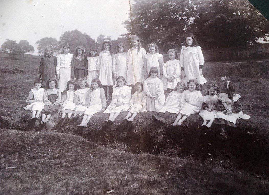 A black-and-white photograph showing a group of school girls outdoors, seated and standing on a grassy bank. They are wearing light-colored dresses typical of the early 1900s. Trees and a stone wall are visible in the background.