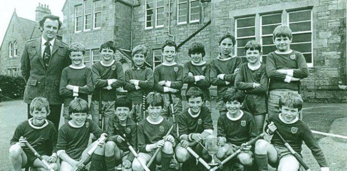 Historic team photo of a school shinty team from the 1980s posed in front of a stone building, wearing matching sports uniforms and holding shinty sticks.