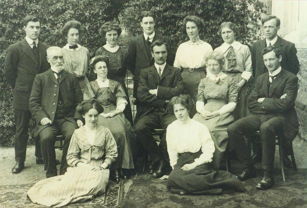 Early 20th-century group photo of male and female teachers from Fort William Higher Grade School posed outdoors in two rows, wearing formal suits and long dresses, with a patterned rug in front.