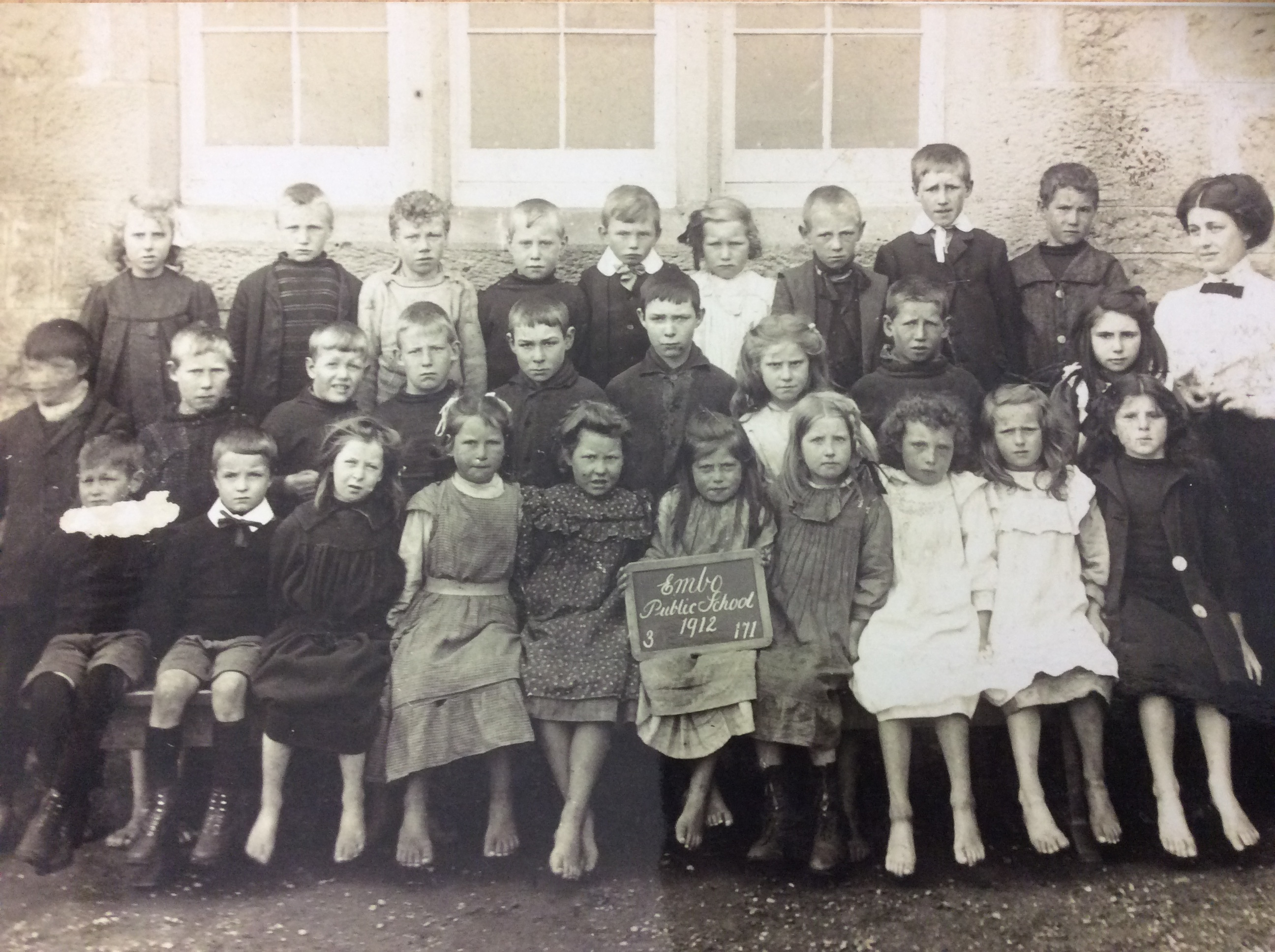 A black-and-white photograph of a large group of schoolchildren posed in three rows outside a stone building with three-paned windows. The children are wearing early 20th-century clothing, including dresses, pinafores, and buttoned jackets. Most are seated on a bench in the front row, with others standing behind. Several children have bare feet, while others wear boots. One child in the front row holds a chalkboard sign that reads: “Embo Public School, 1912. 3 171."