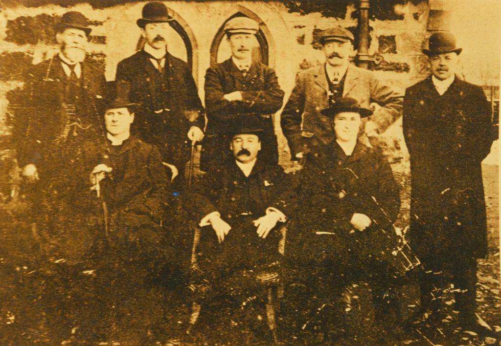 Sepia-toned historic photograph of seven individuals posed in two rows outside a stone building with arched windows. The group is dressed in formal late 19th-century attire, including dark suits, waistcoats, and bowler hats. Two seated figures hold walking sticks.