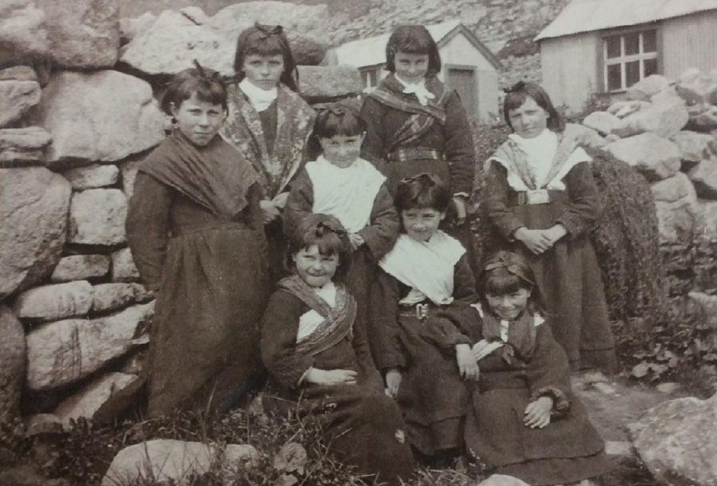 Historic black-and-white photograph showing a group of seven schoolgirls posed outdoors against a large stone wall with two buildings in the background. They are wearing heavy garments with shawls and head coverings, and some have white cloths draped across their shoulders. Taken on St Kilda.