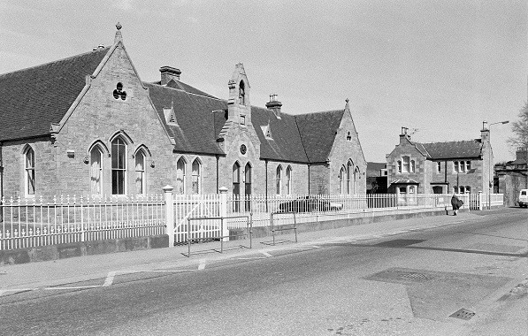 A black-and-white photo of Merkinch Primary School taken from the street in 1986. The image shows a row of stone buildings with steep gabled roofs, arched windows, and a central entrance. A white metal fence runs along the front, and a paved road is in the foreground.