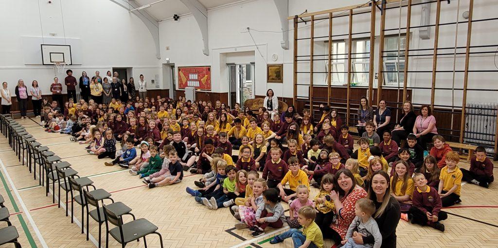 Large group of pupils in maroon and yellow uniforms seated on a gym floor with staff standing at the back; chairs line the left side of the hall.