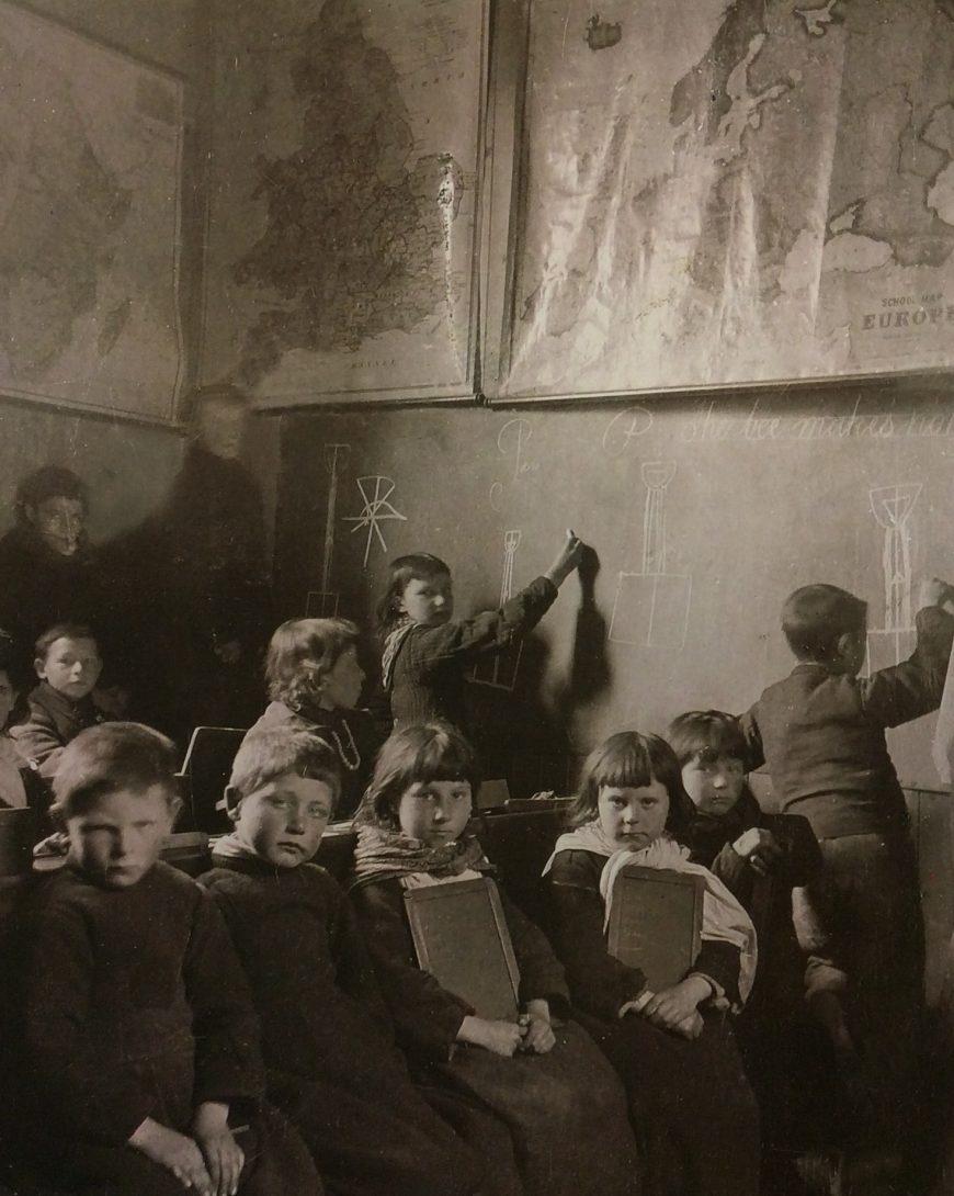 Historic classroom scene with several children seated and holding slates, while a few stand at a large chalkboard drawing geometric shapes. Two large maps, one of Britain and one of Europe, are displayed on the wall above the chalkboard.